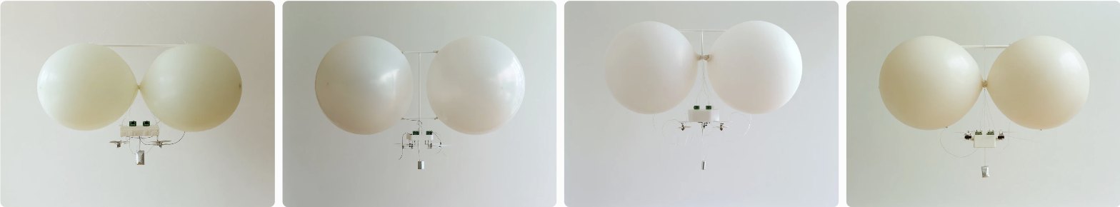 Render: two pale latex balloons in a light yoke with a small white tray of electronics hung beneath, in a plain room